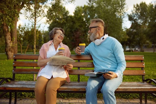 Happy loving couple spending time and leisure activity together on pension in park. Elderly man and woman spouses reading newspaper and book, drinking coffee and sharing positive emotion