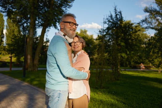 Smiling senior couple hugging with love feeling bonding and gratitude to life giving chance to be happy on retirement. Public green park city outdoors background