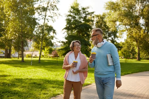 Happy fashioned elderly couple having coffee break walking in park. Romantic aged mature man and woman looking happy enjoying healthy lifestyle on pension