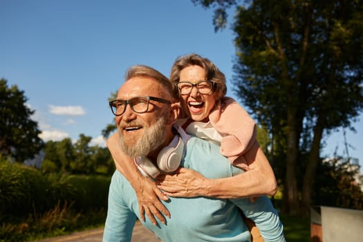 Happy active senior couple having fun outdoor in city park. Portrait of elderly man husband giving piggyback to woman wife