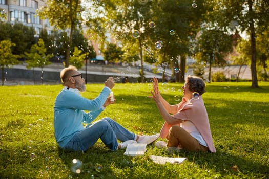 Playful active senior couple blowing soap bubbles while rest in park. Vitality and active elderly people concept