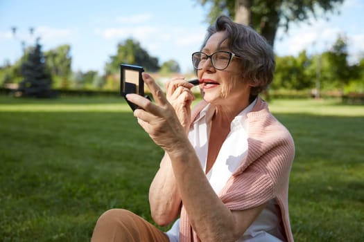 Senior woman applying lipstick doing makeup for charming smile in compact mirror while resting outdoor in city park