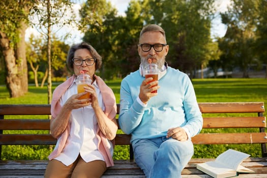 Portrait of relaxed elderly couple drinking refreshment cocktail in takeaway cup while sitting on bench in park. Happy retired life