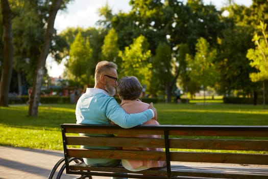 Happy couple enjoying tranquility and idyllic atmosphere in public park while sitting on bench. View from back on cheerful loving man and woman having romantic date