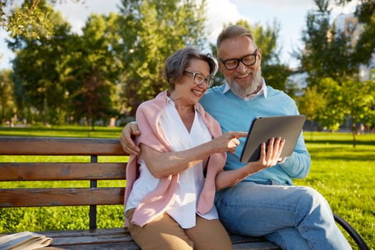 Happy cheerful senior couple using digital tablet while rest on park bench. Modern elderly lifestyles concept