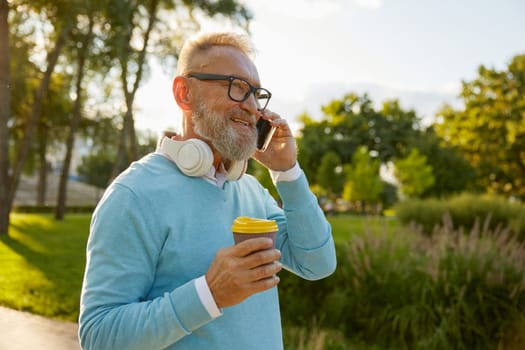 Side view portrait of senior man having call on smartphone. Elderly businessman walking in park enjoying coffee break and talking with relatives on cellphone
