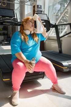 Tired plus size woman wiping sweat from her face with towel feeling exhaustion after cardio training on treadmill machine