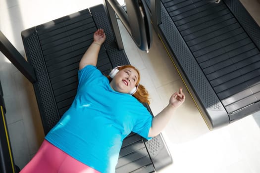 High angle view on happy but tired overweight woman lying on treadmill machine track. Sport, training at gym and obesity concept