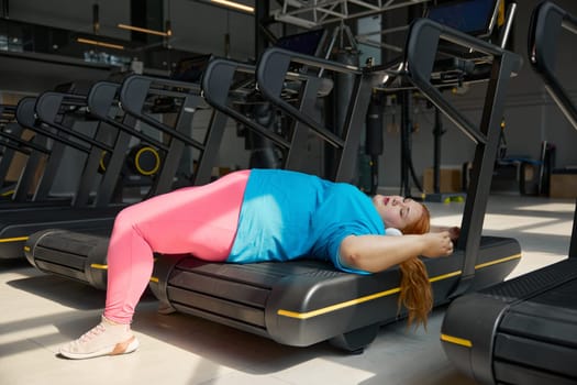 Tired overweight woman lying on treadmill machine track. Sport, training at gym and obesity concept