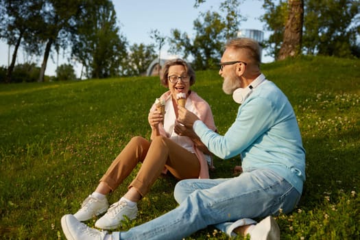 Happy joyful senior couple eating ice-cream in park. Elderly married man and woman fooling around making jokes having snack during walk