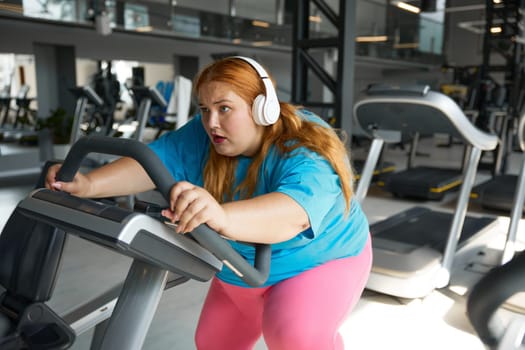 Crazy motivated overweight woman riding fast on stationary bike in gym. Fitness class for obese people concept