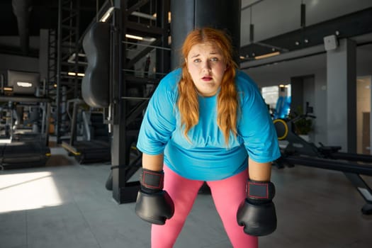 Portrait of pretty young woman with excess weight in boxing gloves feeling tired. Intense effective cardio training for slimming