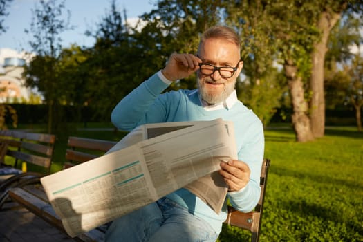 Confident bearded mature man reading daily newspaper on park bench. Morning pastime on retirement