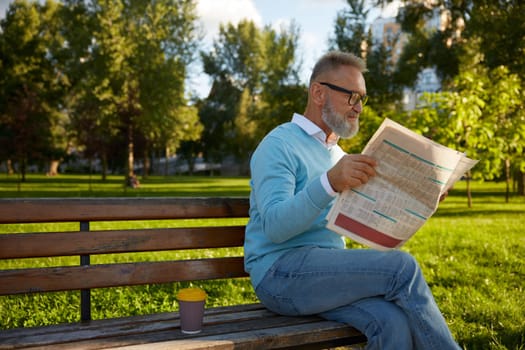 Serious bearded mature man reading daily newspaper on park bench. Morning pastime on retirement