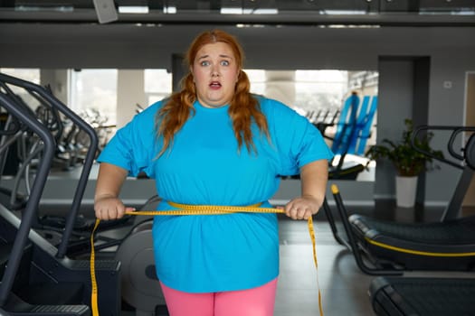 Portrait cute woman with excess weight ties herself with measuring tape standing at gym. Slimming diet and sport training workout for obese female