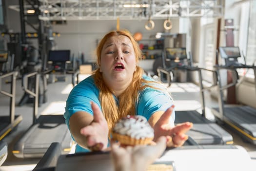 Closeup front view on face of obese woman at gym reaching to cake with hands. Choice between sport for slimming and unhealthy food with obesity