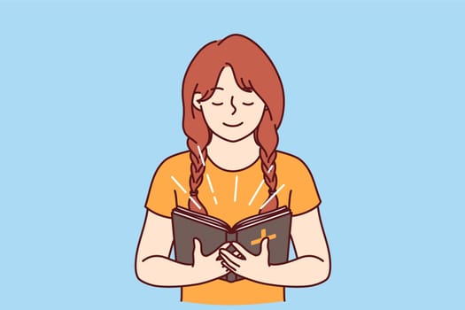 Little girl with bible in hands studies prayers and gospel before going to christian church. Happy child believer in catholic religion holds bible illuminating face and smiles feeling support of lord