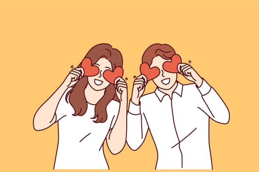 Romantic couple of man and woman holds valentine cards in form of hearts in front of eyes and looks at screen smiling. Romantic guy and girl in white clothes rejoicing in presence of soulmate