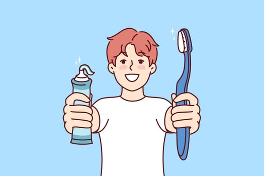 Little boy holds toothpaste and toothbrush showing snow-white smile and recommending brushing teeth regularly. Child independently monitors health of own teeth in order to get rid of caries