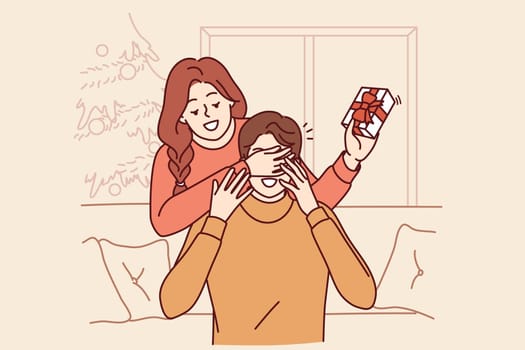 Woman gives christmas gift to boyfriend sitting on sofa and closes eyes to make surprise. Man and wife are in house with christmas tree and exchange new year gifts in beautiful packaging.