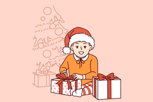 Little pre-teen boy sits near christmas tree and gifts from santa claus and smiling looks at screen. Kid rejoices at arrival of christmas or new year and large number of giftboxes from parents