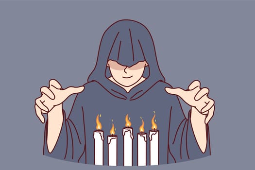 Man monk in hood stands over burning candles, performing mysterious religious ritual to communicate with otherworldly world. Monk experiments with black magic and practices witchcraft