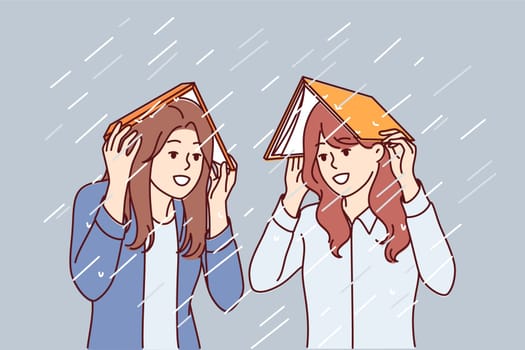 Women take shelter from rain under books and smile, rejoicing at onset of autumn season. Happy girls cover heads with textbooks to avoid getting wet during heavy rain or spring storm