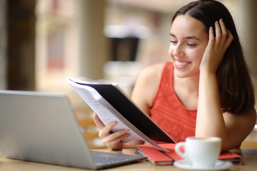 Happy student comparing notes with laptop in a coffee shop terrace