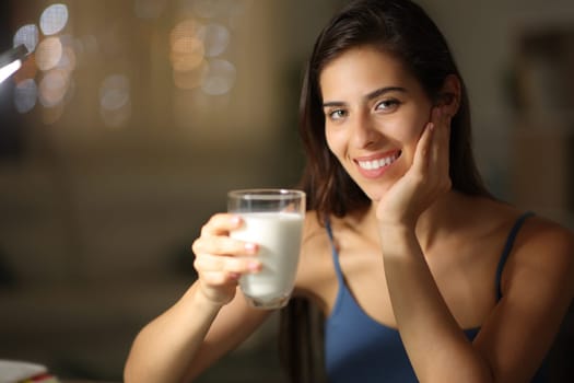Happy woman in the night holding milk glass at home looking at you