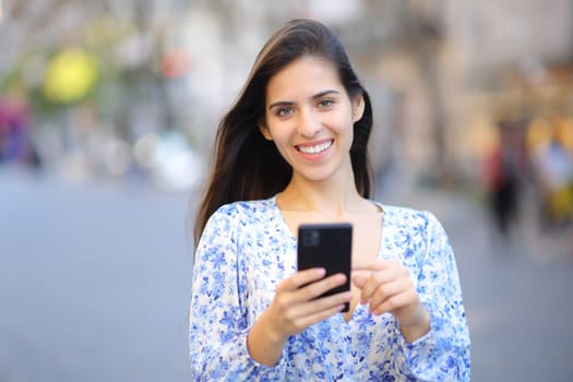 Front view portrait of a happy woman in the street looking at camera using phone