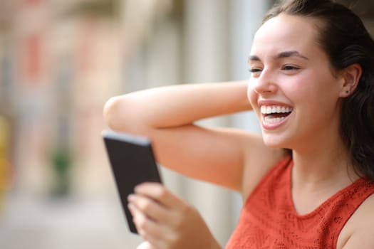 Happy woman laughing holding smartphone looking away in the street
