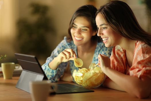 Two happy friends watching movie on tablet eating potato chips in the night at home