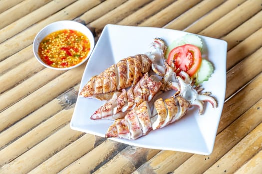 3 grilled squid sit on clean white plate on bamboo table alongside slices of tomato and cucumber. There is a bowl of spicy seafood dipping sauce next to it. It's fresh seafood in a seaside restaurant.