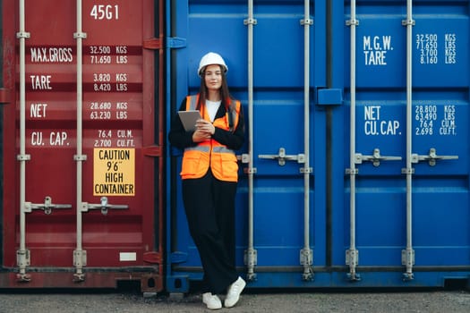 Female Engineer Containers Terminal Worker with tablet posing at container. White helmet and vest in container terminal port or construction site.