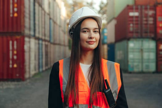 Portrait of beautiful engineer woman with walkie-talkie in white helmet and vest working in container terminal port.