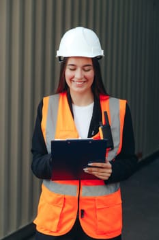 Female Engineer with Clip Folder in white helmet and vest working in container terminal port or construction site.