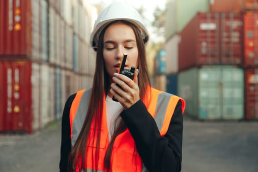 Woman engineer speak to walkie-talkie at shipping containers terminal. Woman in white helmet and vest working.