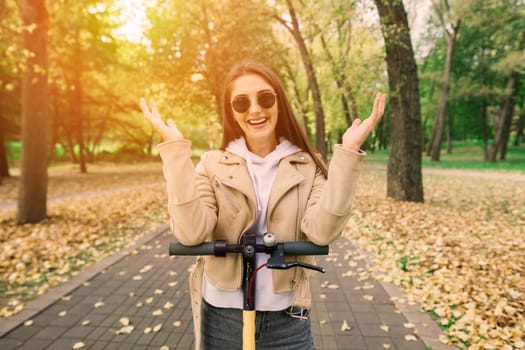 Young beautiful stylish woman riding electric scooter in autumn park, ecological transport