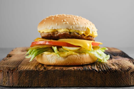 Beef cheeseburger with vegetables and cheesy sauce on wooden board, front view