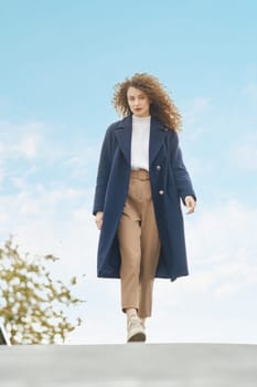 Young woman in blue wool coat walks on the bridge in sunny autumn day