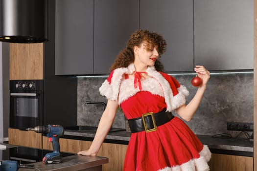 Christmas gift concept - new kitchen furniture. Cheerful woman in santa dress looks at toy ball.
