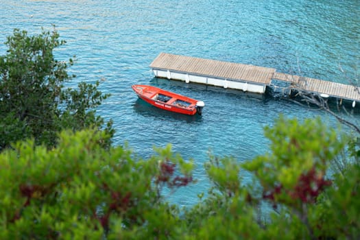 View to calm sea bay from above through some coniferous trees, red rescue boat docked near small wooden pier.