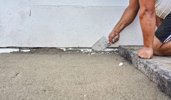 Installing new pavement or floor outside from large concrete tiles, closeup detail on male worker fixing sand and gravel base layer near wall.