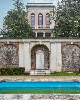 A beautiful white marble drinking fountain located in the garden of the Mecidiye Pavilion, Istanbul, Turkey, with a decorative fountain in front, and the palace in the background