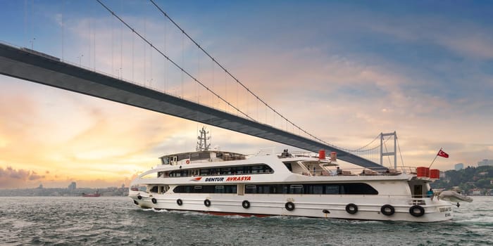Istanbul, Turkey - May 17 2023: White ferryboat sails under the Bosphorus Suspension Bridge before sunset, with the city skyline in the background. Photo taken from Beylerbeyi Palace