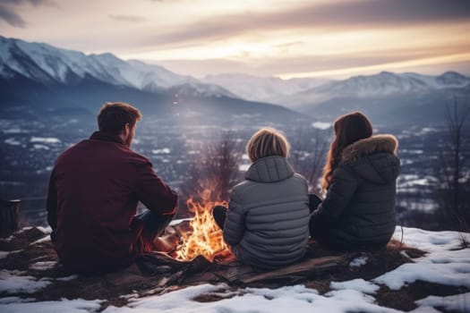 Friends sitting around campfire in evening during mountain winter trip. Winter season. People having fun sitting at bonfire looking at mountain landscape. Enjoying fire in the evening. Weekend trip. Travel to the mountains. Natural destinations