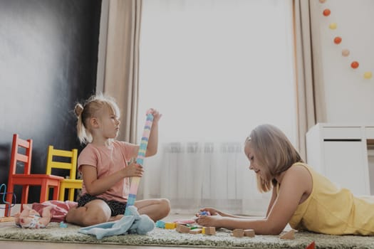Children playing with colorful toys. Kids sitting on carpet at home bedroom or kindergarten, nursery. Educational eco-friendly game for child. Adorable little girls build house, tower together