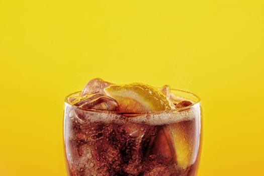 glass filled with Coca-Cola, ice cubes, and a slice of lemon. Close up of the upper part on bright yellow background.