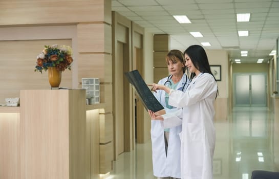 Two women doctor working together examining patient x-ray scan in hospital . Senior doctor and young smiling doctor standing in hallway and discussing diagnosis of brain x ray results. Healthcare, medicine and insurance concept