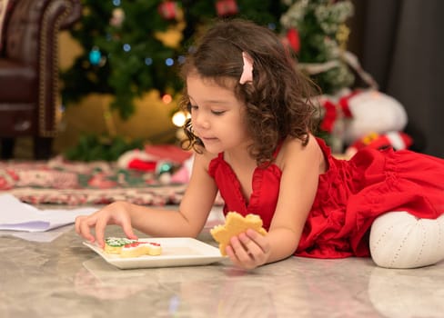 Cheerful toddler girl in red dress holding cookies and enjoying sweets while lying on floor in front of decorated Christmas tree on Xmas eve. Kid celebrating Christmas at home.